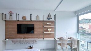 A modern living room with a flat-screen TV mounted on a wooden wall, a dining area with a table and chairs, and a balcony with a view of the city.
