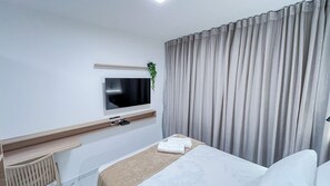Een moderne hotelkamer met een flatscreen-tv aan de muur, een nachtkastje met een afstandsbediening, een bed met witte lakens en een groot raam met grijze gordijnen.