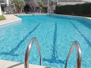 A clear swimming pool with metal ladder steps, surrounded by a landscaped area with palm trees and a hedge. - Les Gavines - Two Bedroom Apartment, Sleeps 4 (L'Escala)