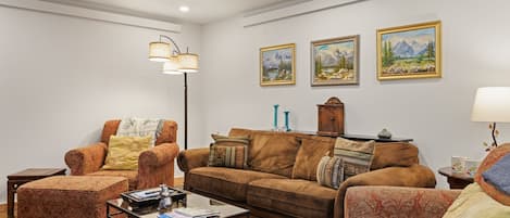 Een woonkamer met een bruine bank, fauteuil en poef, een salontafel en een vloerlamp. Er hangen drie ingelijste schilderijen aan de muur.