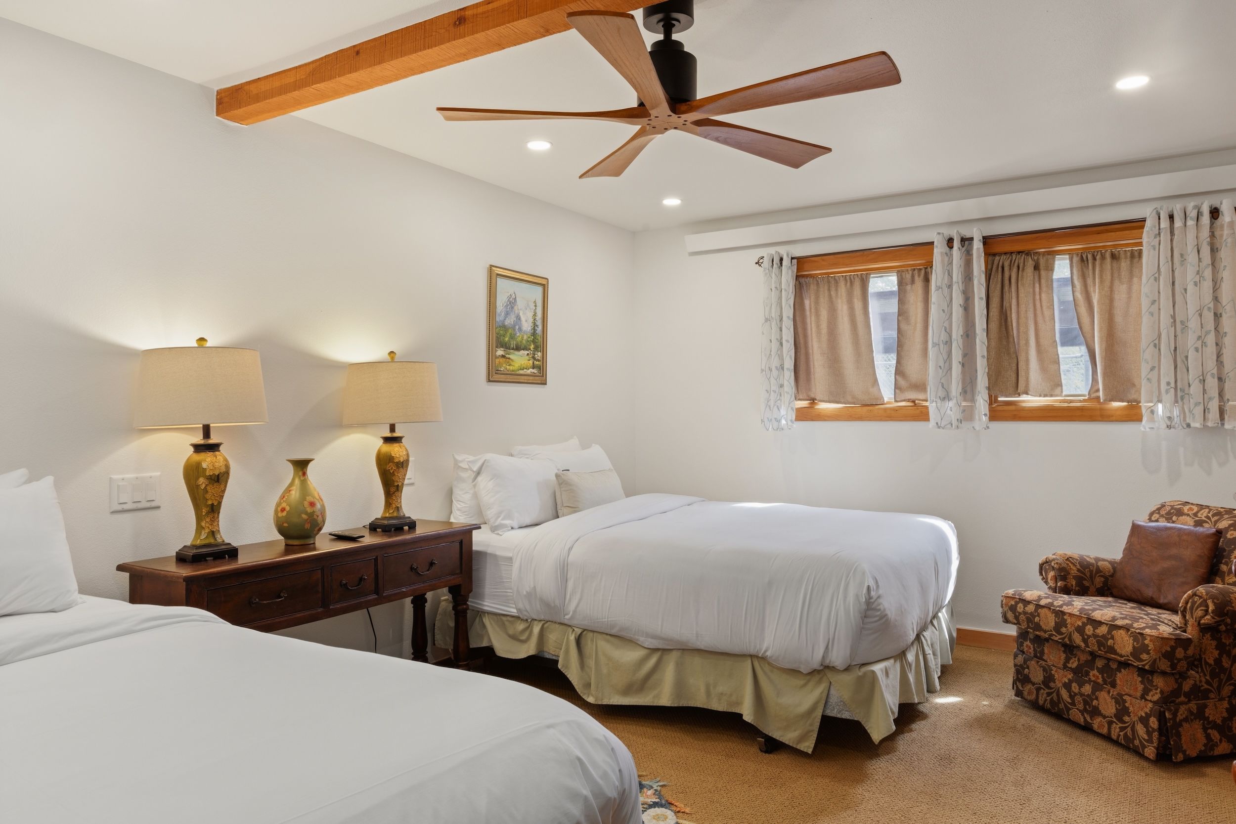 Quarto com duas camas, ventilador de teto, mesa de cabeceira com abajures, uma cadeira e janela com cortinas.