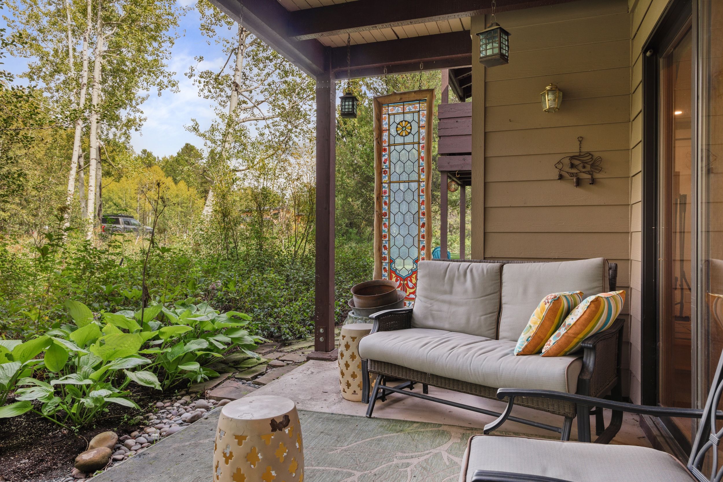 Un patio cubierto con una zona de estar acolchada, una puerta decorativa con vidrieras y vistas a un área boscosa.