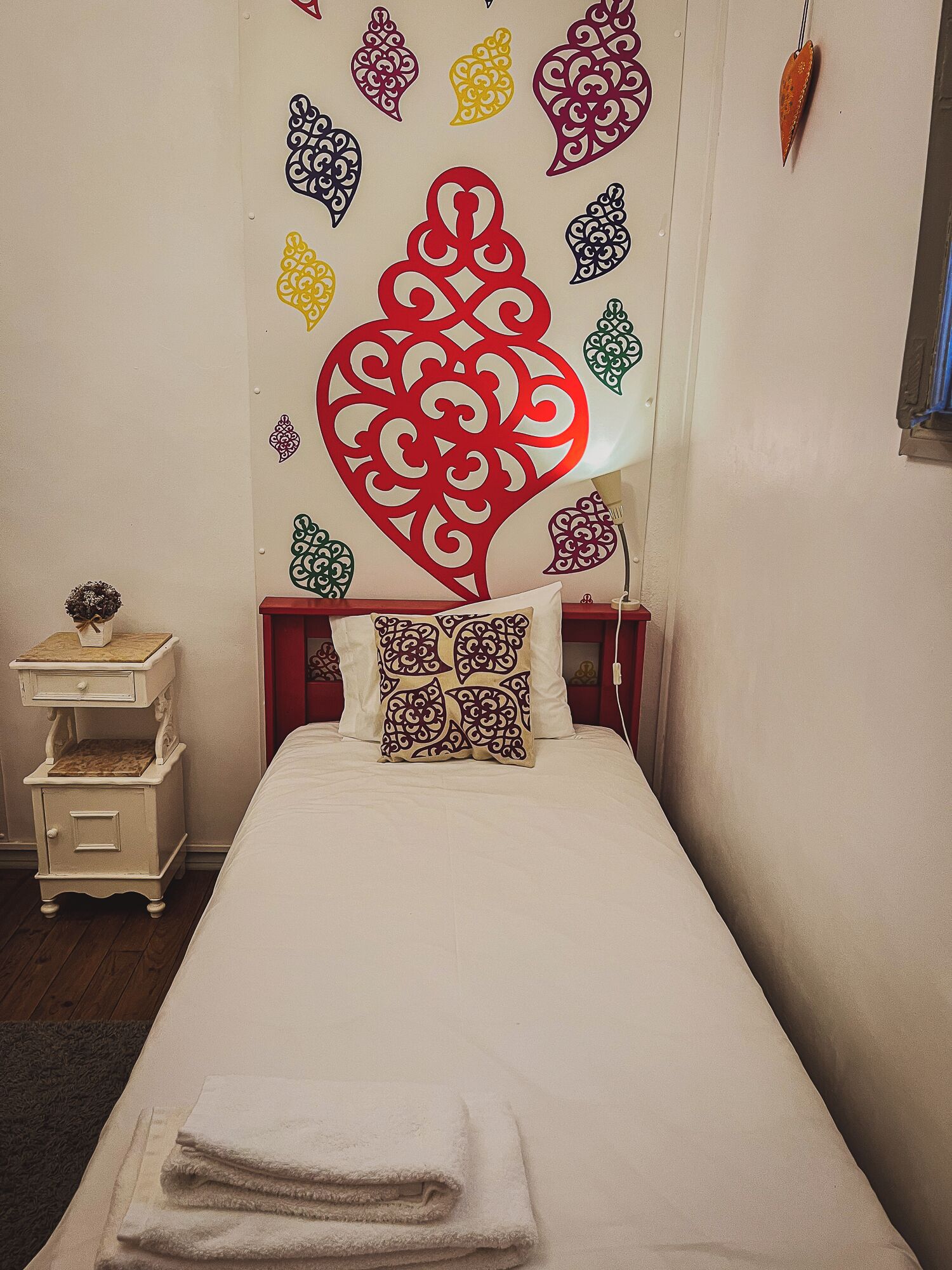 A single bed with a white bedspread and two pillows, a nightstand with a vase, and a wall with decorative heart patterns.