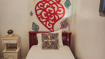A single bed with a white bedspread and two pillows, a nightstand with a vase, and a wall with decorative heart patterns.
