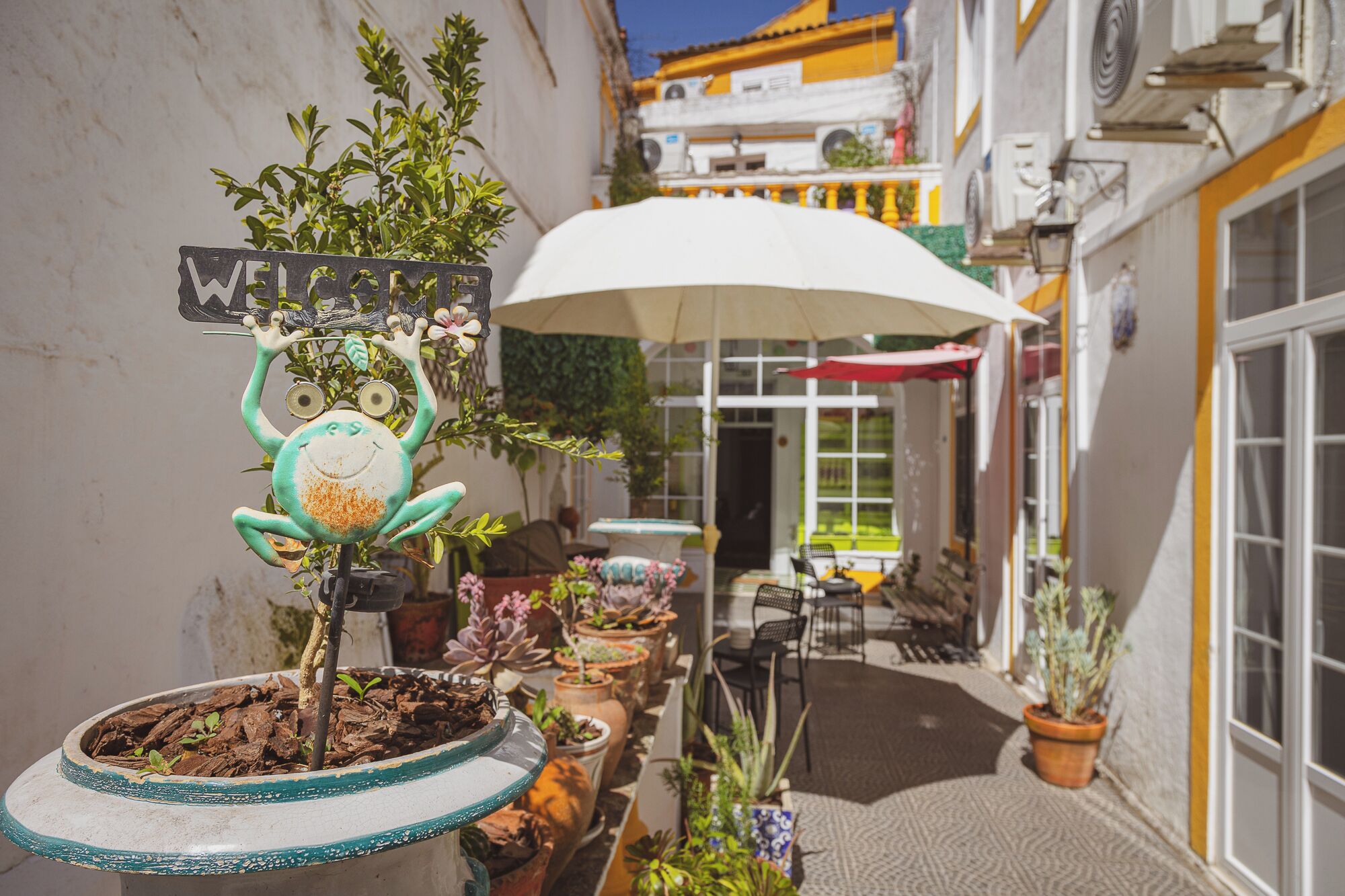 A narrow outdoor corridor with potted plants, a whimsical 'WELCOME' sign, and a white umbrella providing shade.