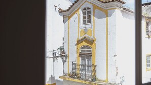 A view through a window of a building with a yellow and white facade, a balcony with a metal railing, and a cobblestone street. - Very Typical - Alojamento Local (Elvas)