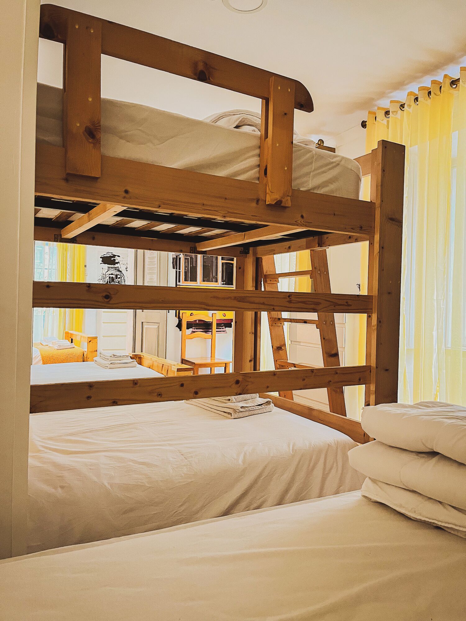 A bunk bed room with wooden beams, a window with yellow curtains, and a small table with a book.