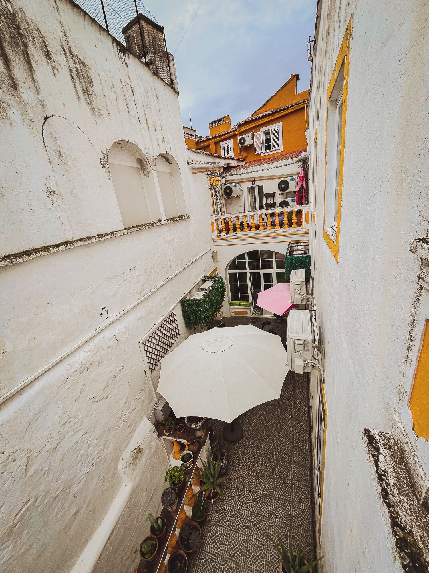 Une ruelle étroite avec une table parasol blanche, des plantes en pot et un sol carrelé.