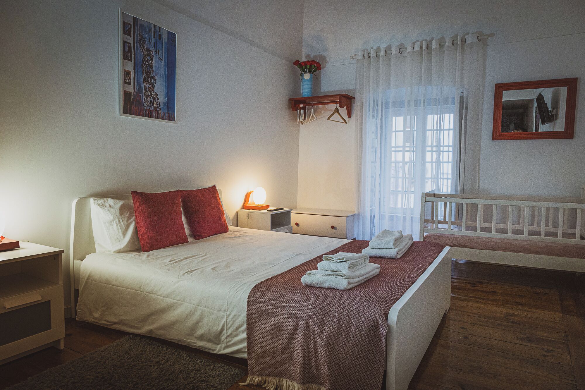 Un dormitorio con cama, mesitas de noche, un tocador, un espejo, una cuna y una ventana con cortinas.