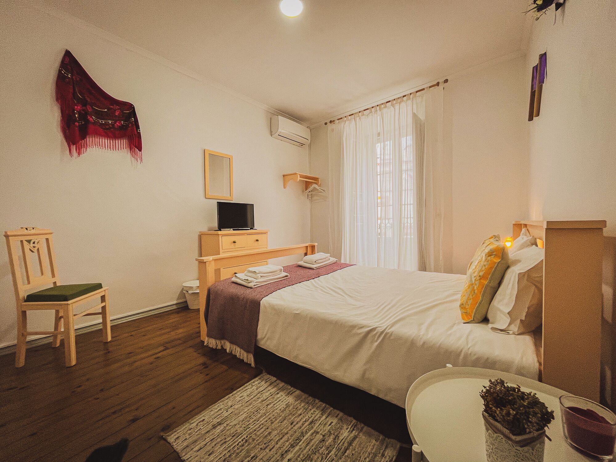 A bedroom with a bed, a chair, a TV, a mirror, and a window with curtains.