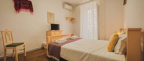 A bedroom with a bed, a chair, a TV, a mirror, and a window with curtains.