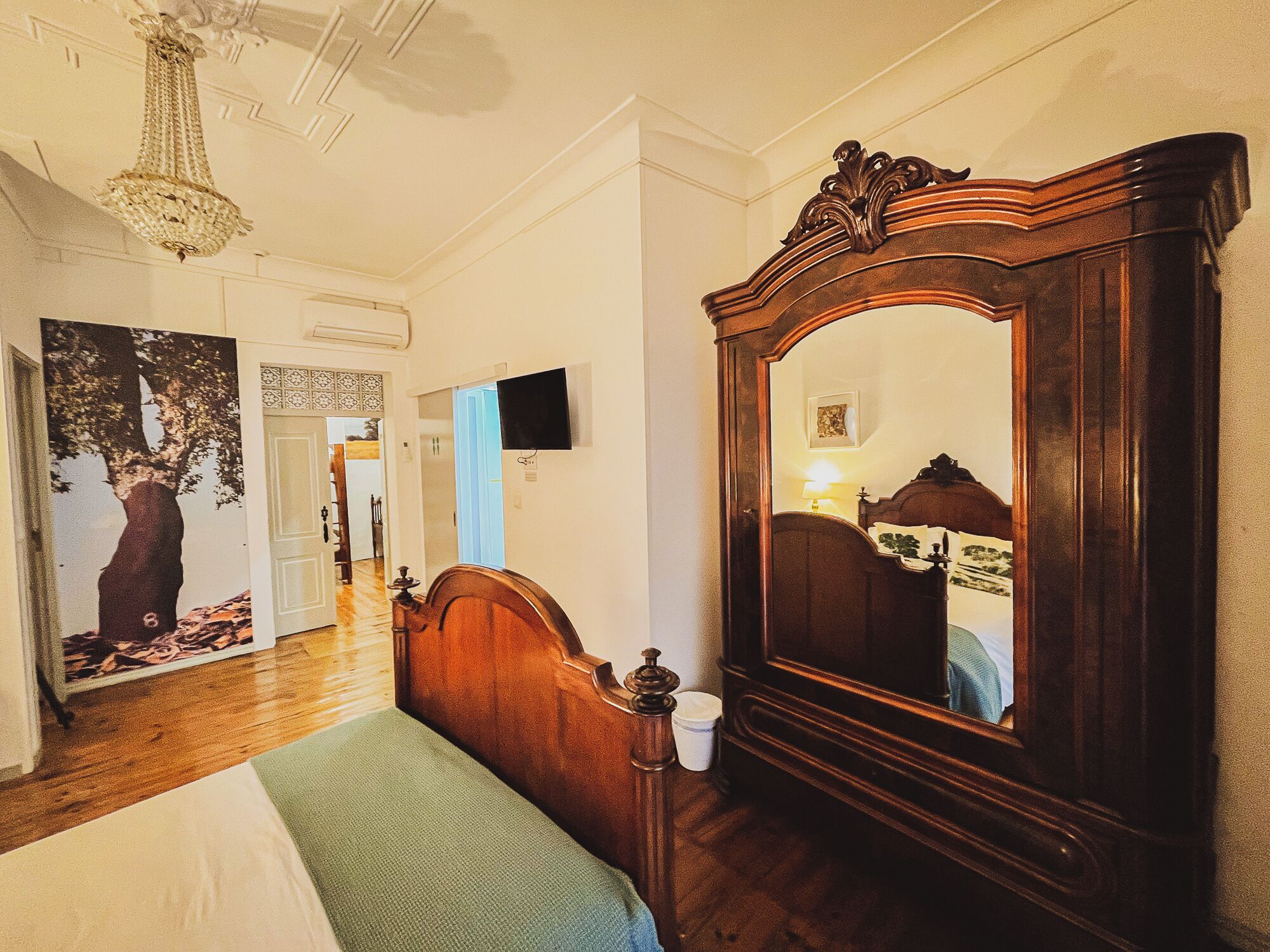 A bedroom with a wooden bed, a large mirror, a chandelier, and a painting of a tree.