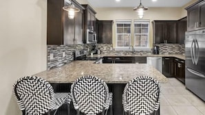 A modern kitchen with dark wood cabinets, a granite countertop, and a stainless steel refrigerator. There are three black and white patterned chairs at the island. - Villa Soleil Dunes (Miramar Beach)