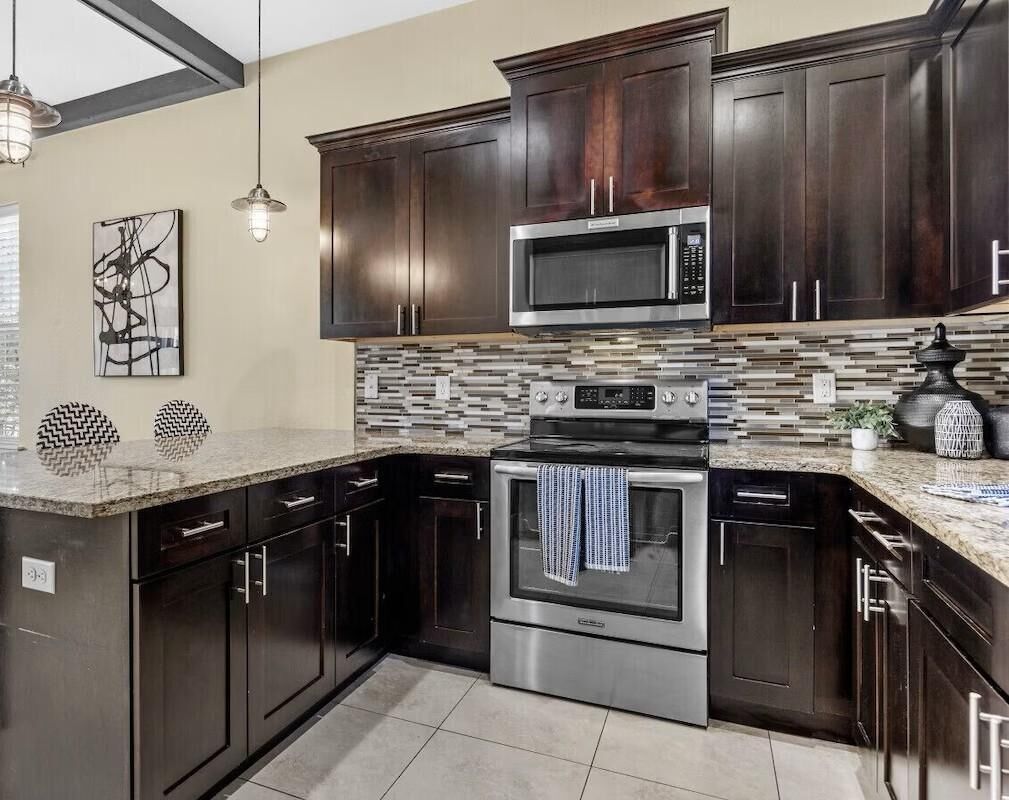 A modern kitchen with dark wood cabinets, a stainless steel microwave, and an oven. There is a tiled backsplash and a granite countertop.