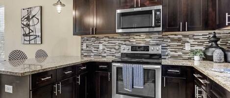 A modern kitchen with dark wood cabinets, a stainless steel microwave, and an oven. There is a tiled backsplash and a granite countertop.