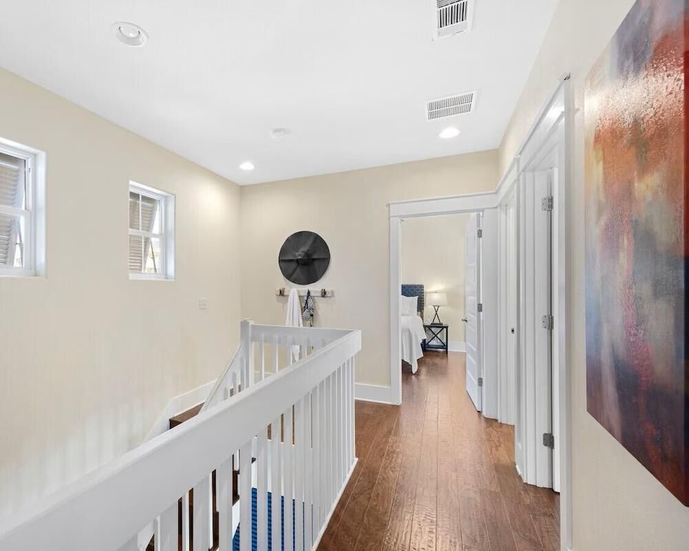 A hallway with a wooden floor, white walls, and a white railing. There is a door at the end of the hallway and a painting on the wall.