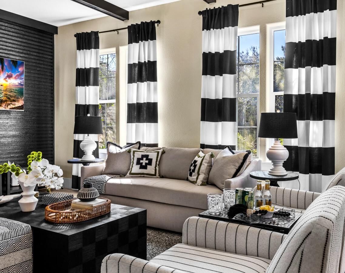 Een moderne woonkamer met een beige bank, gestreept fauteuils en een salontafel. Zwart-wit gordijnen kleden de ramen.
