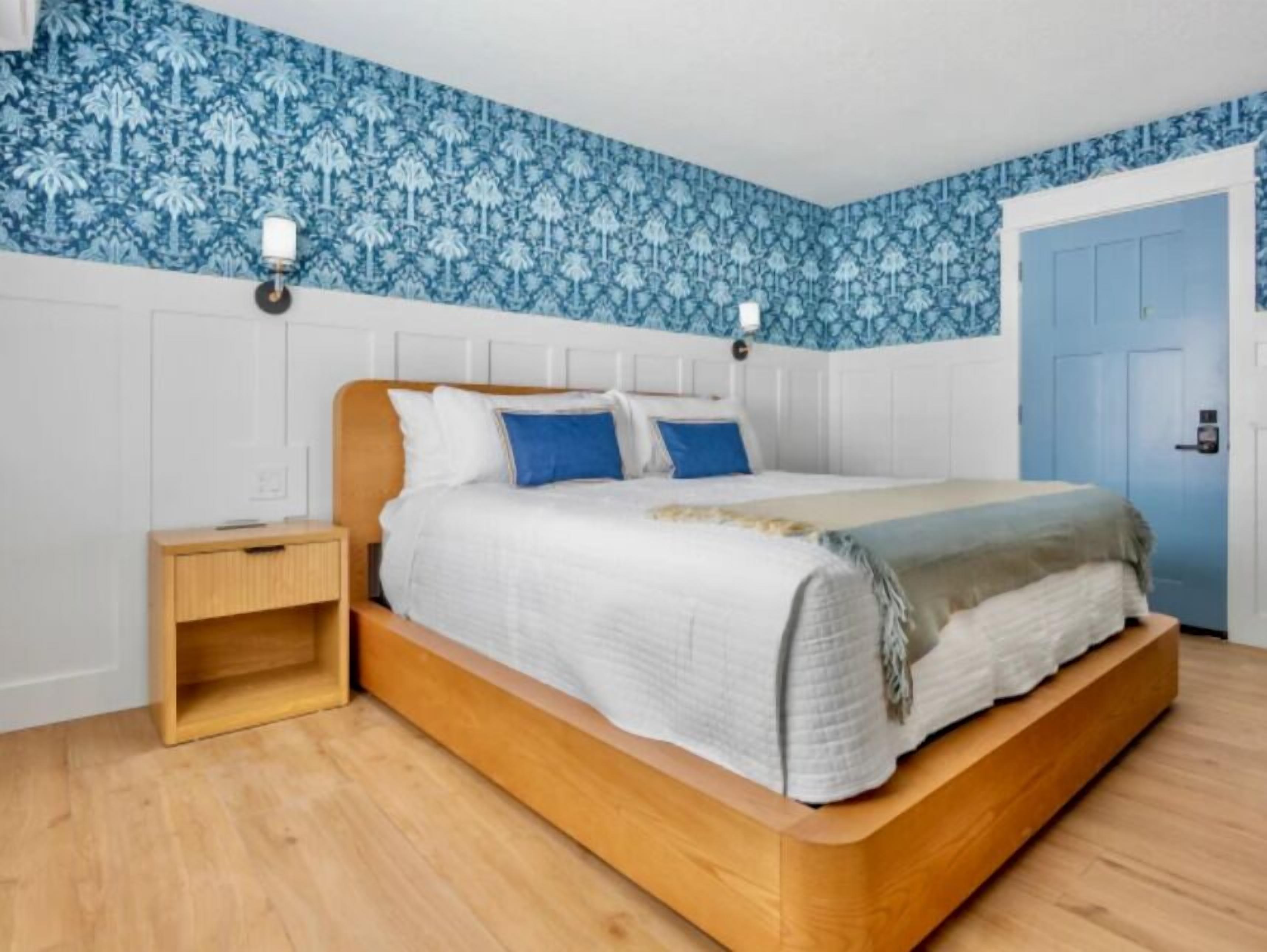 A bedroom with a wooden bed, white bedding, a nightstand, and blue patterned wallpaper.