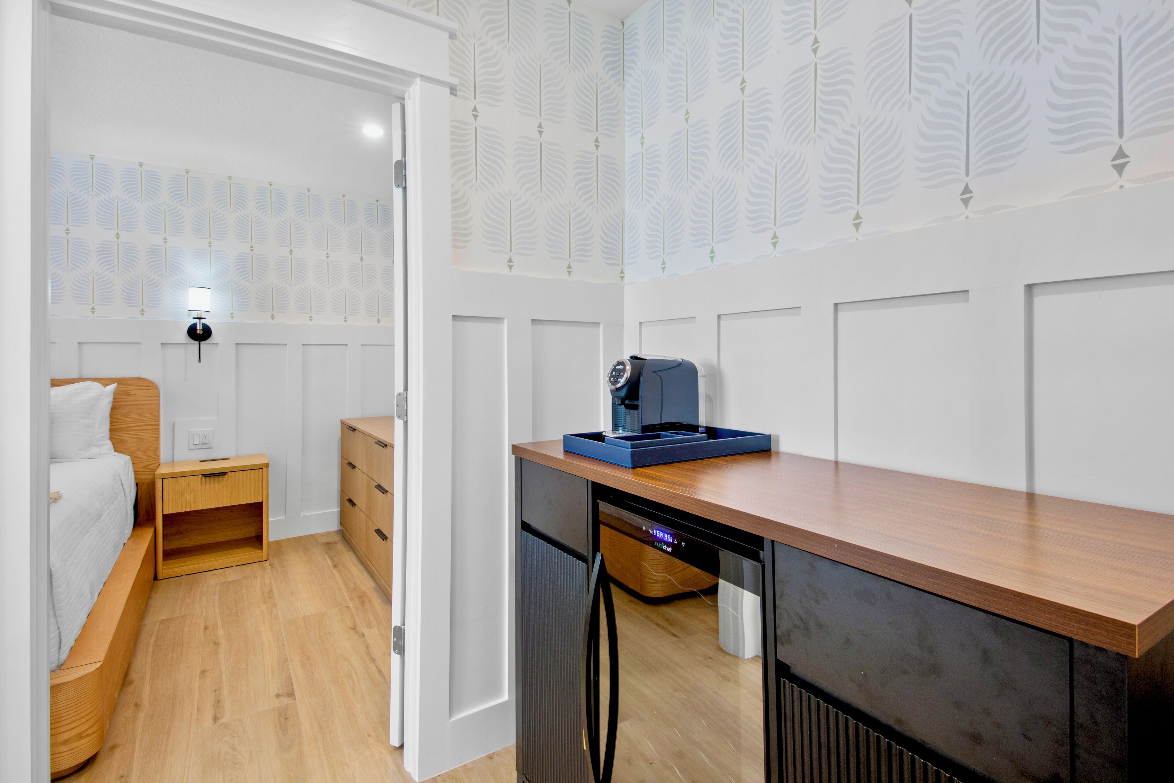 Ein modernes Zimmer mit Schreibtisch aus Holz, integriertem Geschirrspüler, kleinem Nachttisch aus Holz, einem Bett mit weißer Bettwäsche und einer Wandleuchte.