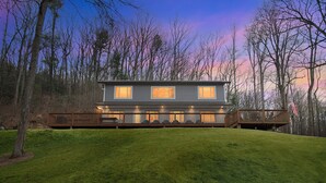 Une maison à deux étages dotée d’une terrasse et d’un porche couvert, entourée d’arbres et d’une pelouse verdoyante.