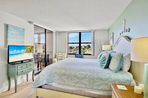 Ein Hotelzimmer mit einem großen Bett, einem Fernseher, einem Schreibtisch mit Schubladen und Blick auf den Strand durch das Fenster.