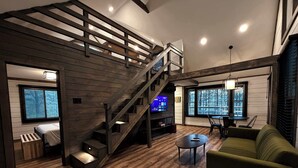 A modern living room with a wooden staircase, a green sofa, a coffee table, and a TV on a wooden shelf. - Hummingbird Hollow • Hot Tub • Private Forest Stay (Irvine)