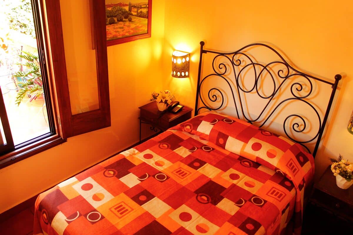 Ein Bett mit gemustertem Überwurf, eine an der Wand befestigte Lampe, ein kleiner Nachttisch mit Telefon und Blumen sowie ein Fenster mit Blick ins Grüne.