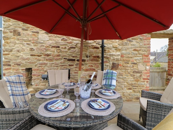 Une table ronde en verre, surmontée d’un parasol rouge, est dressée avec des assiettes, des verres et des couverts.