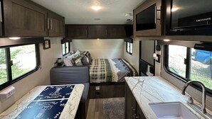 Ein kompaktes Wohnmobil-Interieur mit einer Küchenzeile, einem Bett mit gemustertem Bettbezug und einem Fenster mit Ausblick.