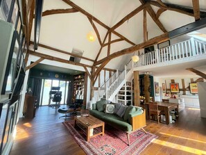 Een ruime loft met zichtbare houten balken, een groene bank, een houten salontafel en een trap naar een hoger niveau.