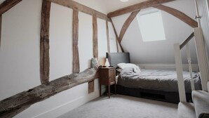 Une chambre avec des poutres apparentes, un lit simple, une table de chevet avec une lampe et une fenêtre laissant entrer la lumière naturelle.
