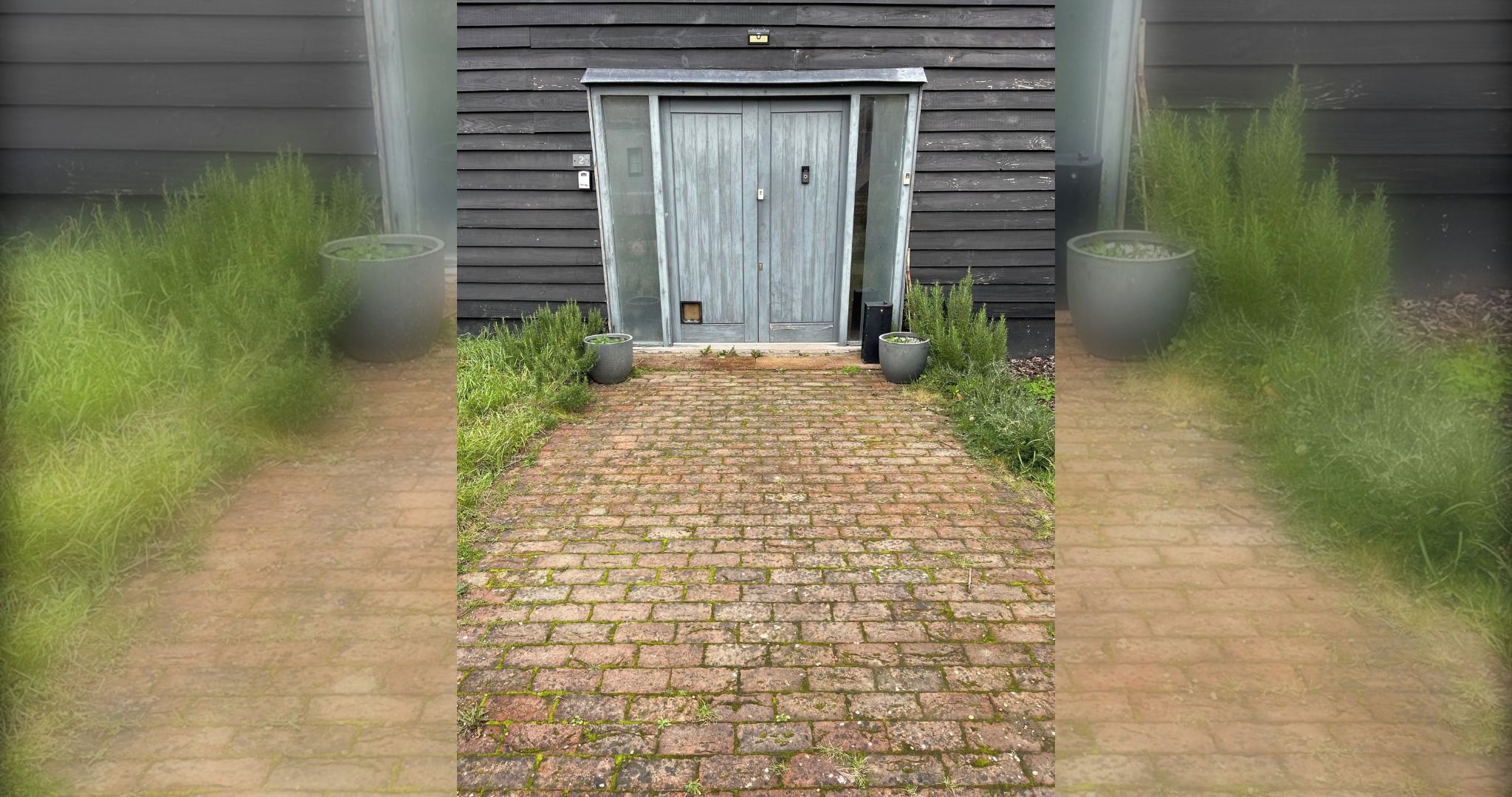A brick pathway leading to a wooden door with a number '2' sign above it, flanked by grass and potted plants.