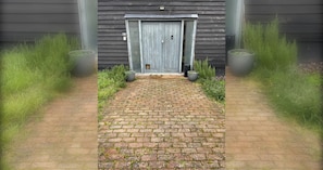 Un camí de maons que condueix a una porta de fusta amb un rètol amb el número '2', flanquejat per gespa i plantes en testos.