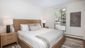 Une chambre moderne avec un lit en bois, deux tables de chevet avec des lampes, une grande fenêtre avec des stores et une œuvre d’art abstraite accrochée au mur.