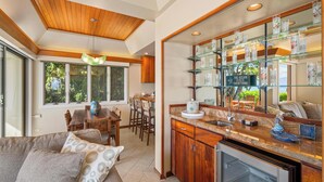 A modern kitchen with a glass display shelf, wooden cabinets, and a dining area with a table and chairs. - Luxury Beachfront Villa: Steps to Baby Beach | Pu'unoa Beach Estates 105 (Lahaina)
