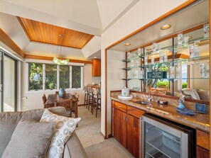 A modern kitchen with a glass display shelf, wooden ceiling, and a dining area with a table and chairs. - Luxury Beachfront Villa: Steps to Baby Beach | Pu'unoa Beach Estates 105 (Lahaina)