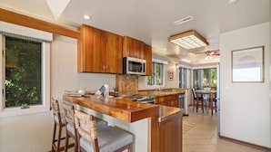 A modern kitchen with wooden cabinets, a central island with bar stools, a microwave, and a dining area with a table and chairs. - Luxury Beachfront Villa: Steps to Baby Beach | Pu'unoa Beach Estates 105 (Lahaina)