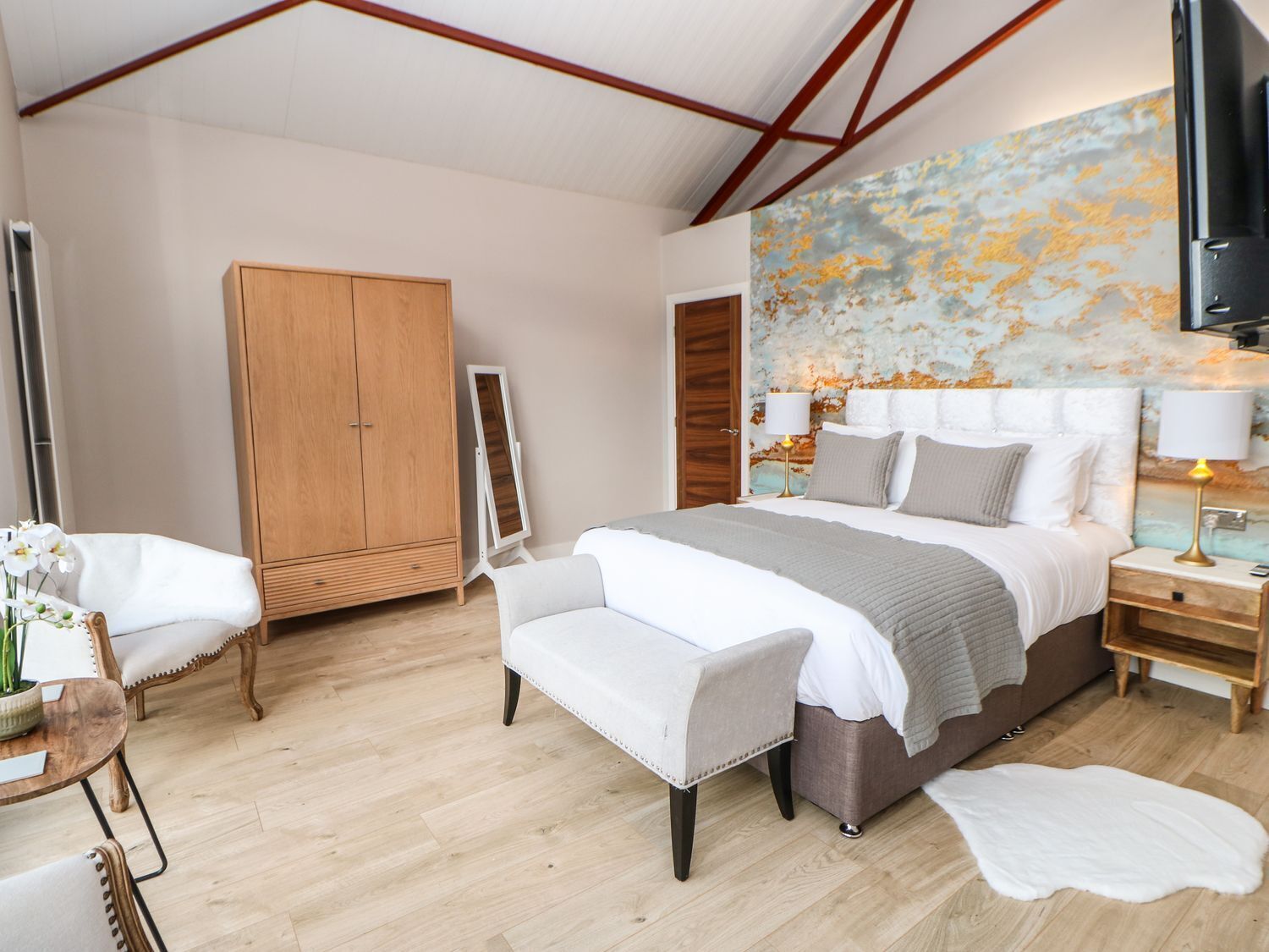 A modern bedroom with a large bed, a wooden wardrobe, a seating area with a chair and ottoman, and a large abstract painting on the wall.