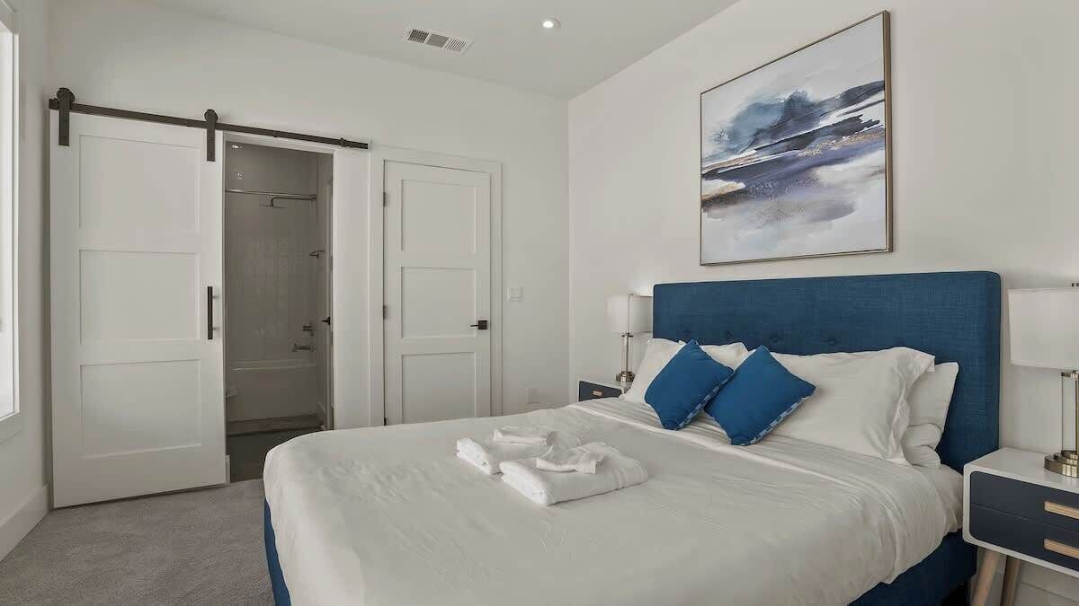 A modern hotel room with a large bed, a blue headboard, white bedding, and a painting on the wall.