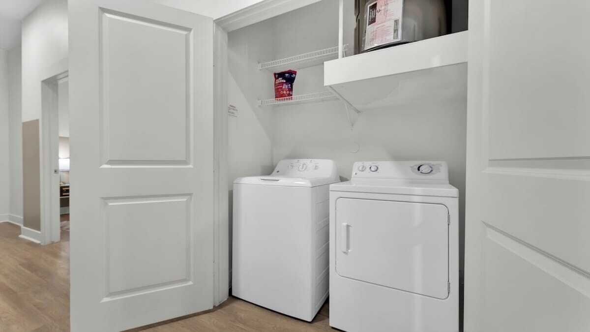 Un cuarto de lavandería con lavadora y secadora, un estante con una caja de detergente y una puerta que da a otra habitación.