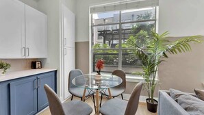 Une cuisine moderne avec des meubles blancs, une table ronde en verre et des chaises grises. On y trouve une plante en pot et une fenêtre donnant sur des bâtiments et de la verdure.