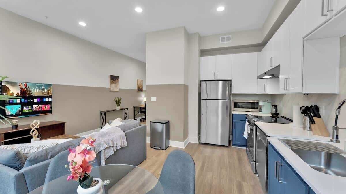 A modern kitchen with stainless steel appliances, a dining area with a gray sofa, and a flat-screen TV mounted on the wall.