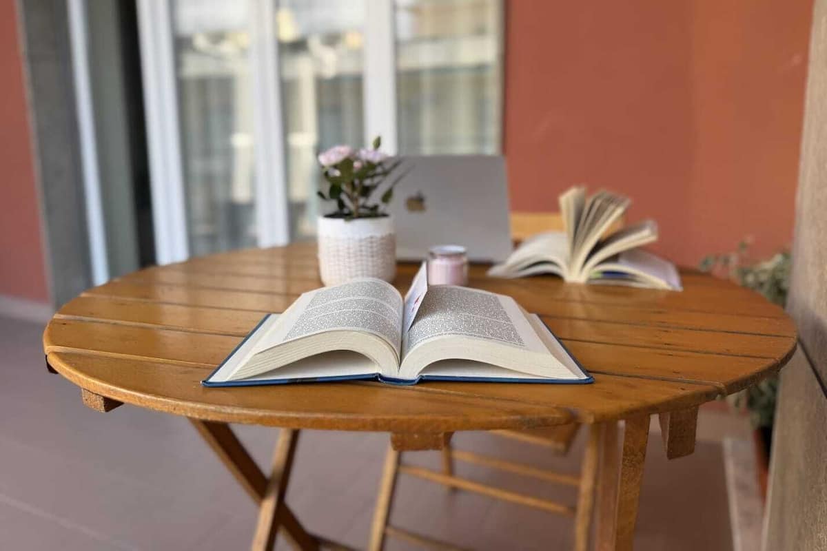 An open book on a wooden table with a laptop and a potted plant in the background.