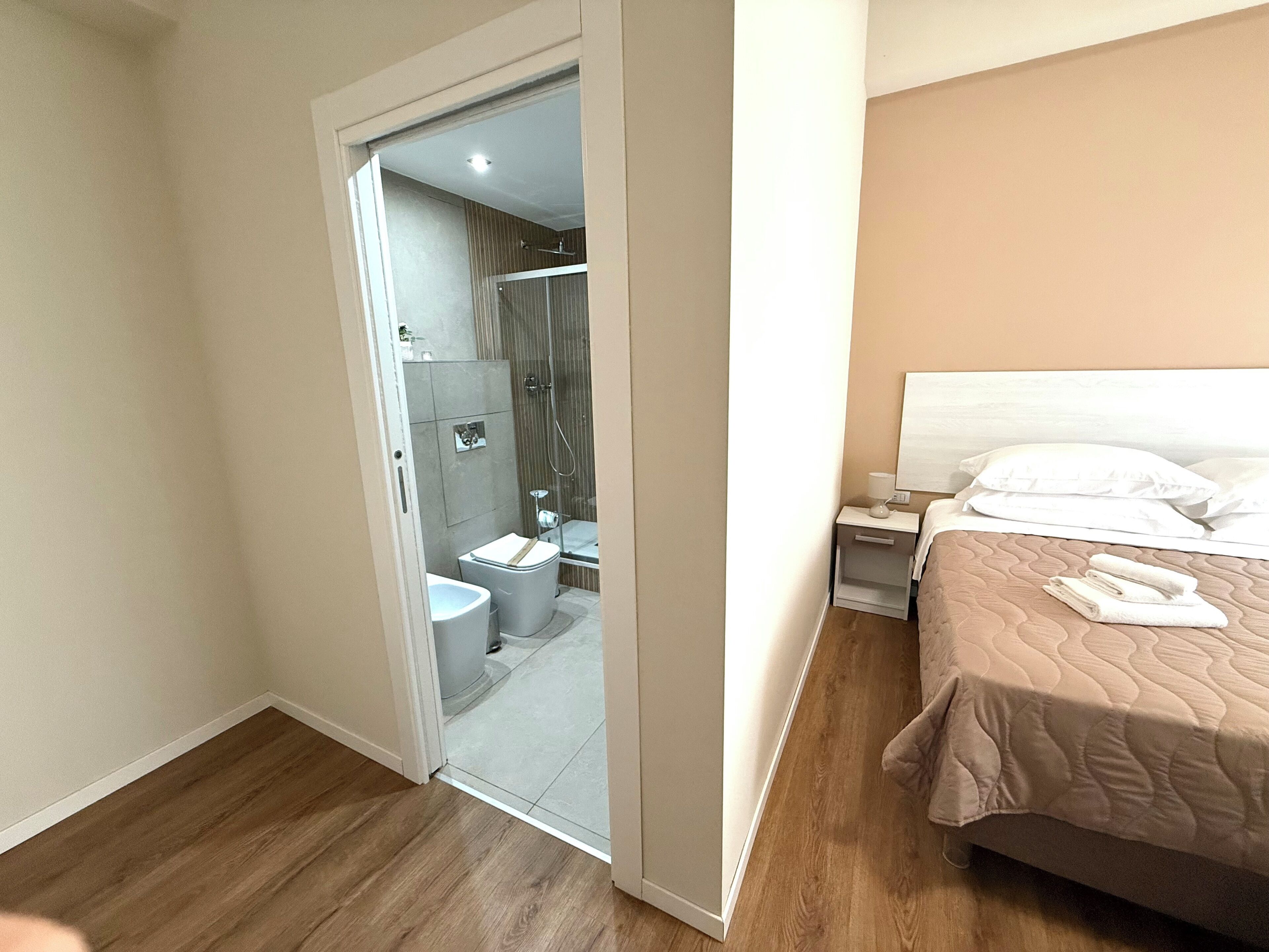 A hotel room with a wooden floor, a bed with a beige quilt, a nightstand with a lamp, and a bathroom with a shower and toilet.