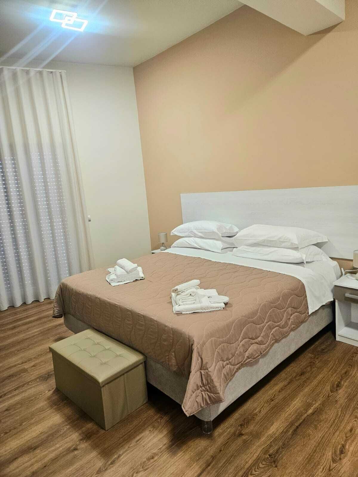 A hotel room with a bed, a nightstand, a bedside table, a window with curtains, and a wooden floor.