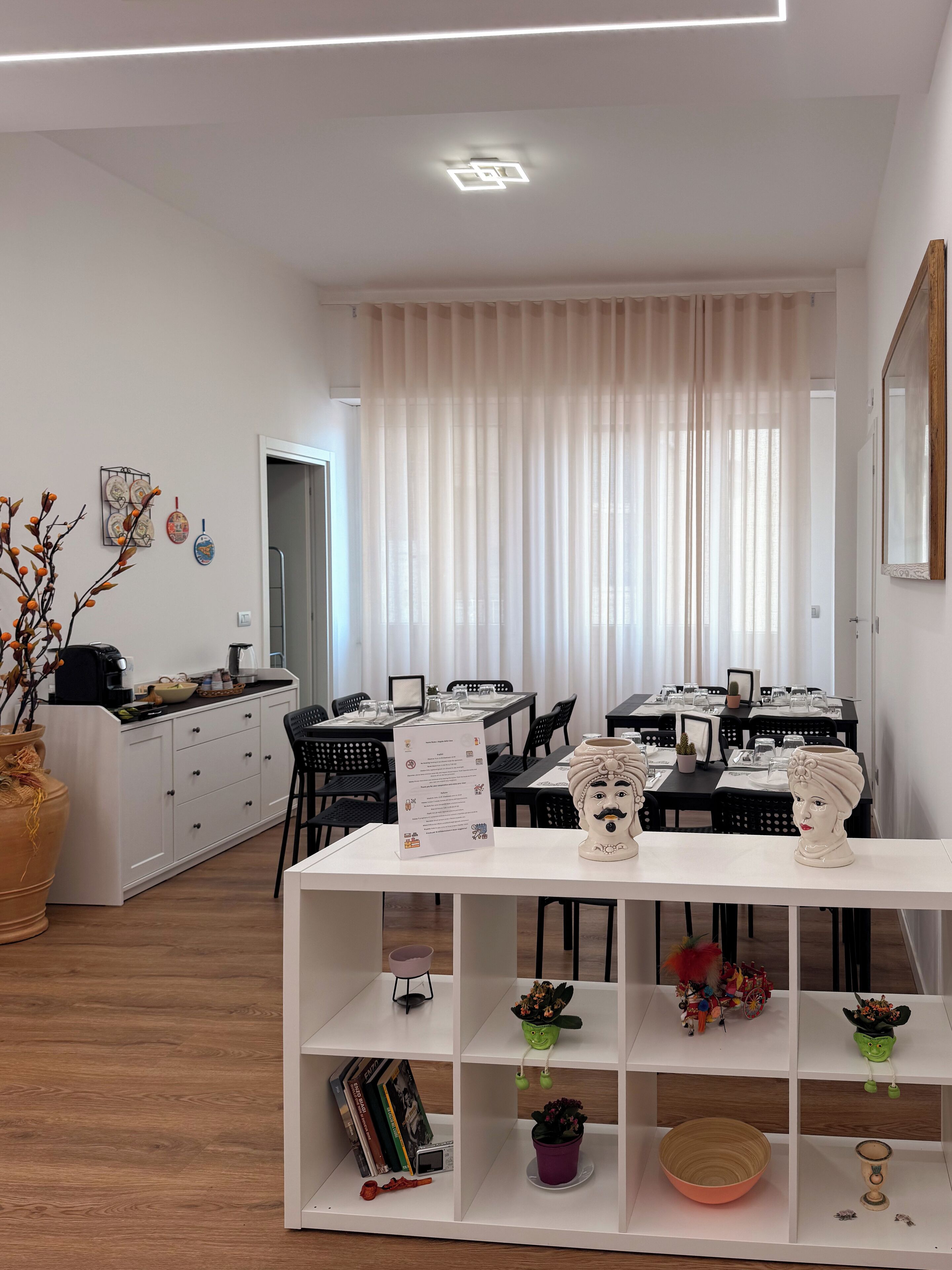 Un comedor moderno con mesas y sillas blancas, un armario blanco con adornos, y un amplio ventanal con cortinas traslúcidas.
