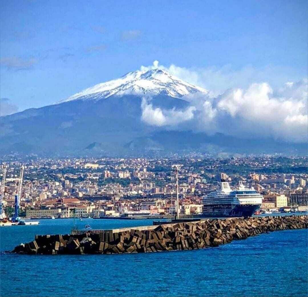 A coastal city with a prominent snow-capped mountain in the background, a cruise ship docked at the harbor, and a rocky breakwater in the foreground.