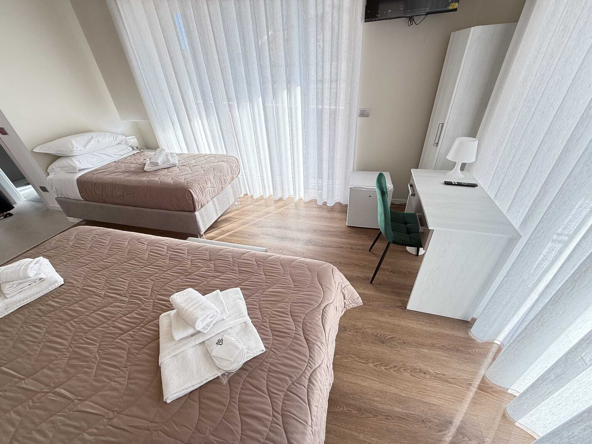 A hotel room with two beds, a desk, a chair, and a large window with sheer curtains.