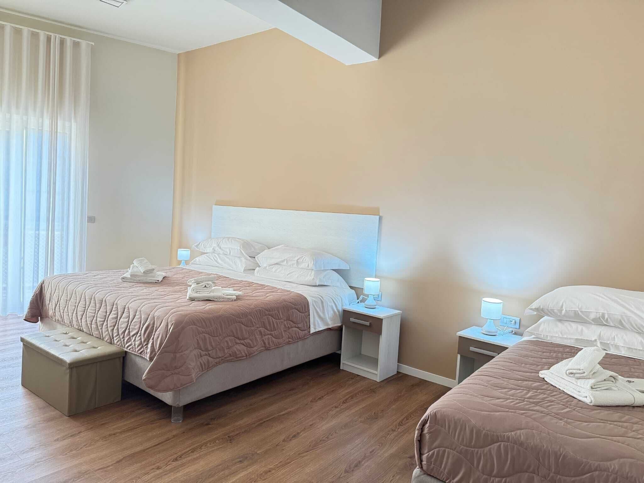 A hotel room with a large bed, a smaller bed, a bench, bedside tables with lamps, and a window with curtains.