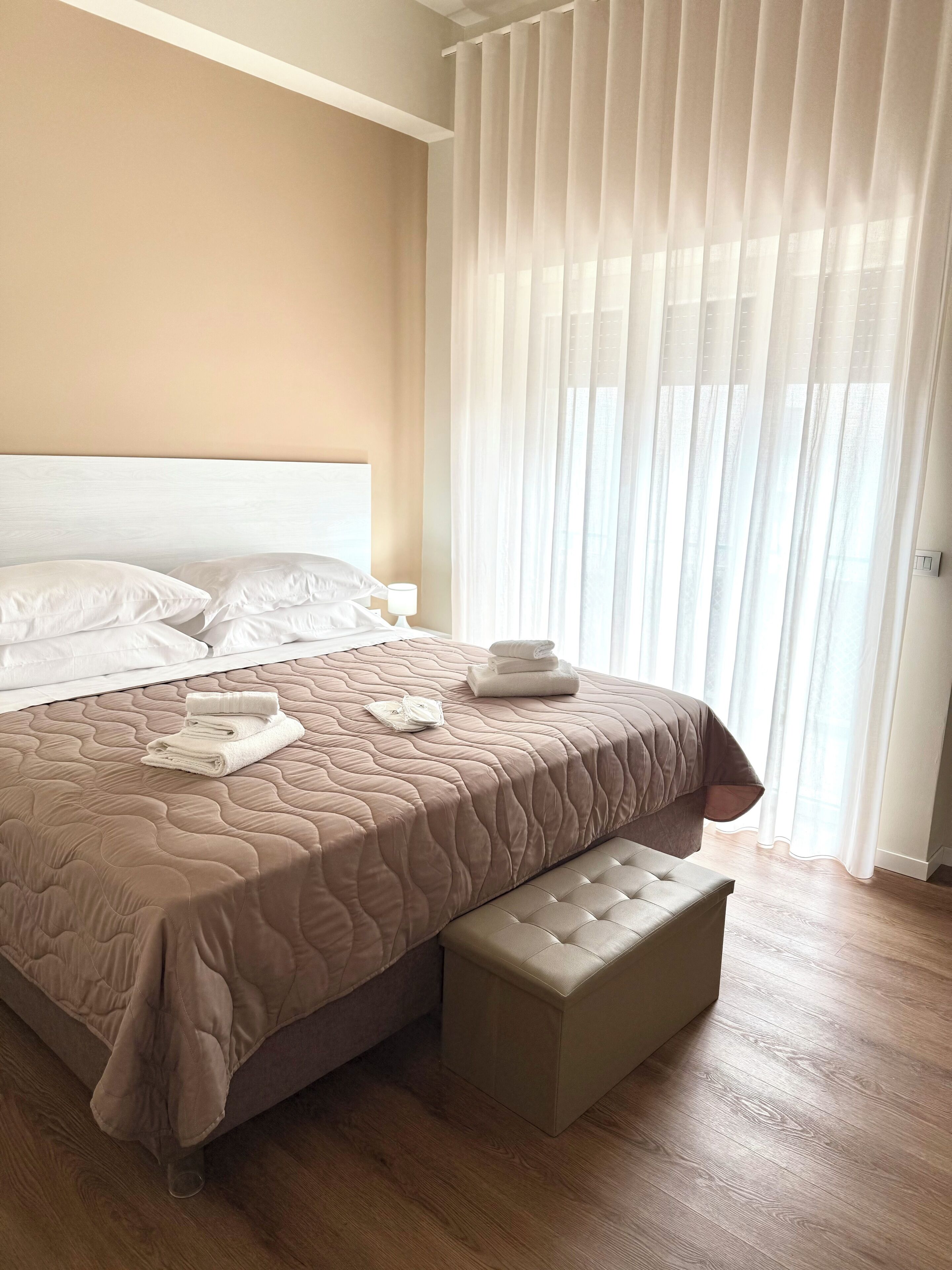 Uma cama bem arrumada com cabeceira acolchoada, mesa de cabeceira com abajur e janela com cortinas leves.